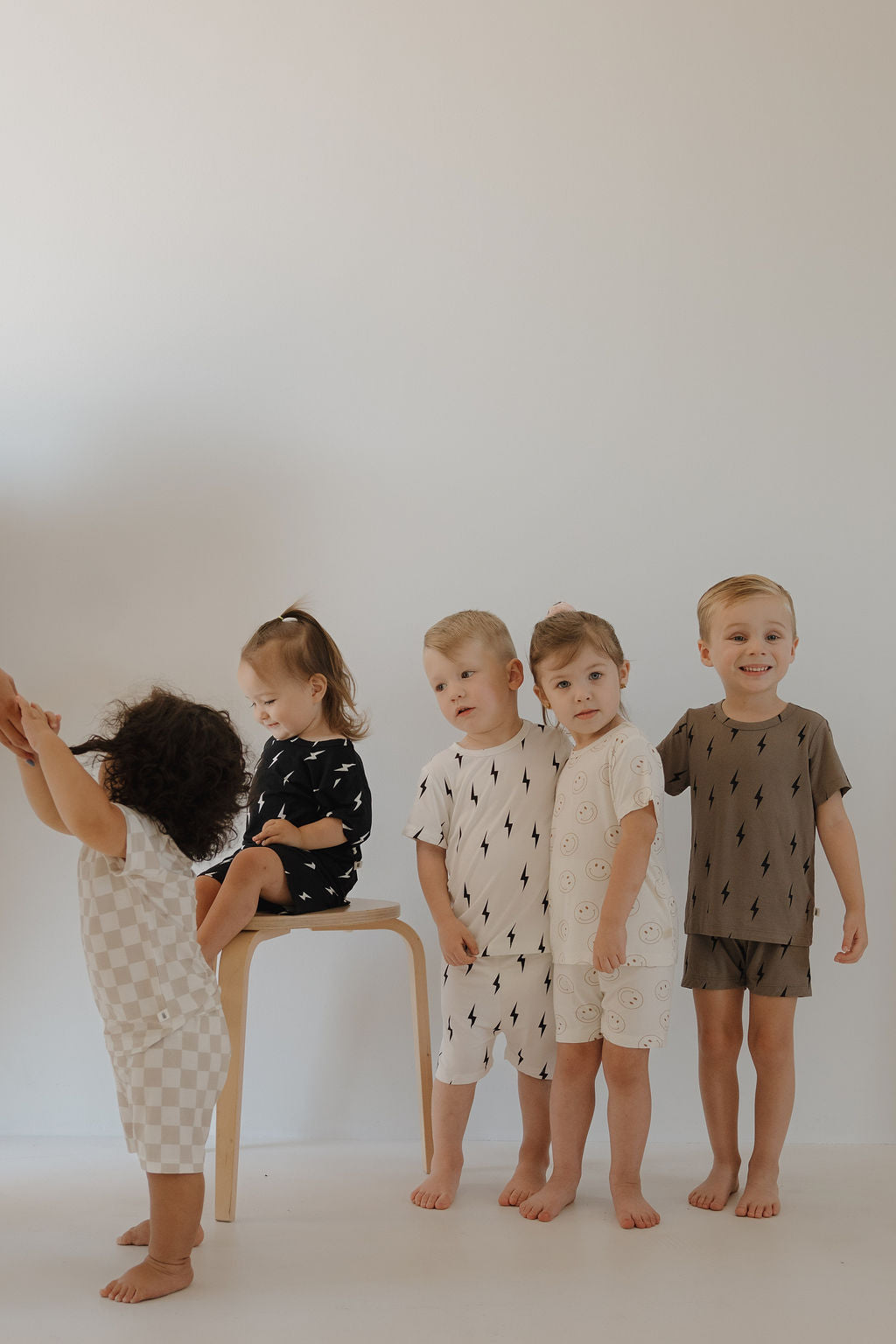 Five young children wear forever french baby Bamboo Short Sets in White & Black Lightning Bolt, posing against a white wall. One stands left, another sits on a stool, and three smile together, barefoot in their breathable pajamas.