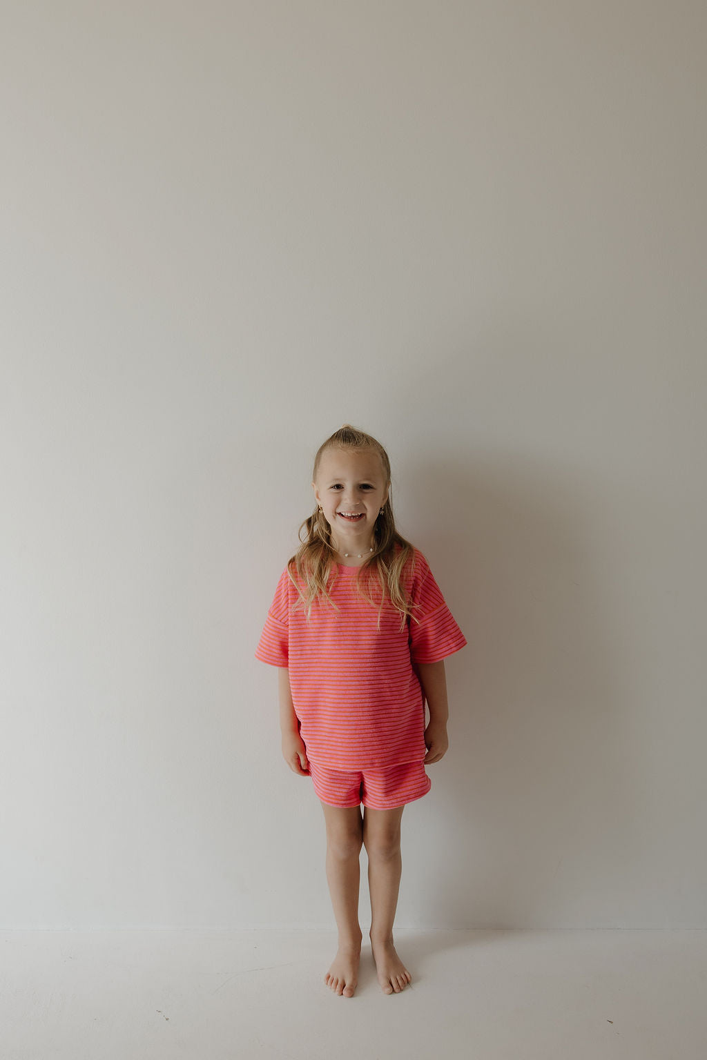 A young child with long blonde hair stands barefoot against a plain white wall, smiling. The child is wearing the forever french baby Child Terry Short Set | Strawberry, ideal for family moments and playful days.