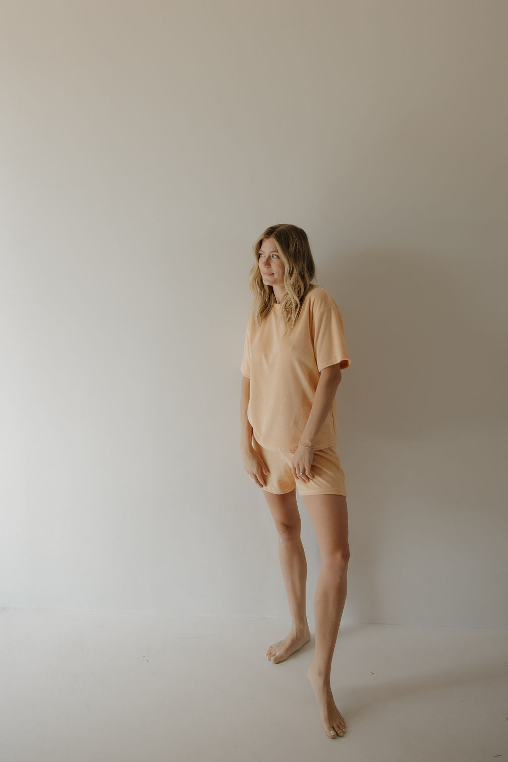 A woman with wavy blonde hair stands barefoot against a light wall, wearing the Women's Terry Short Set in Sherbert from forever french baby, looking to the side and smiling slightly for an effortlessly stylish look.