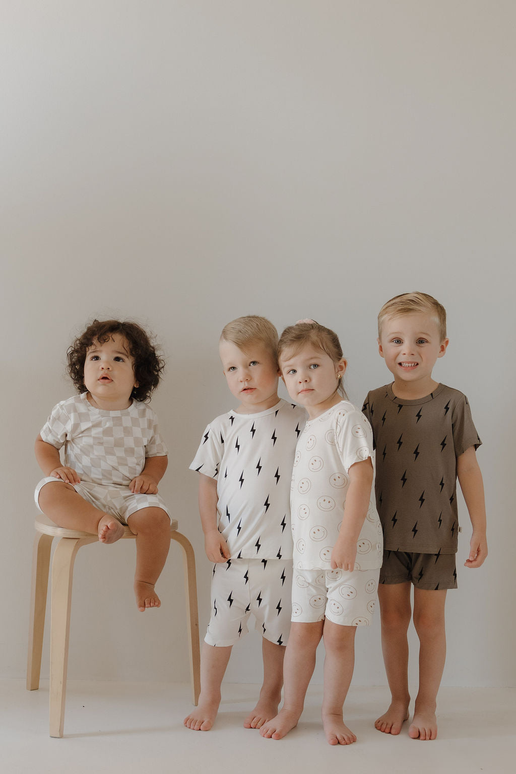 Four young children pose indoors against a plain background. Three stand close together, and one sits on a stool. All wear soft, breathable Bamboo Short Sets in Checkerboard by forever french baby, looking comfy and relaxed with bare feet and gentle smiles.