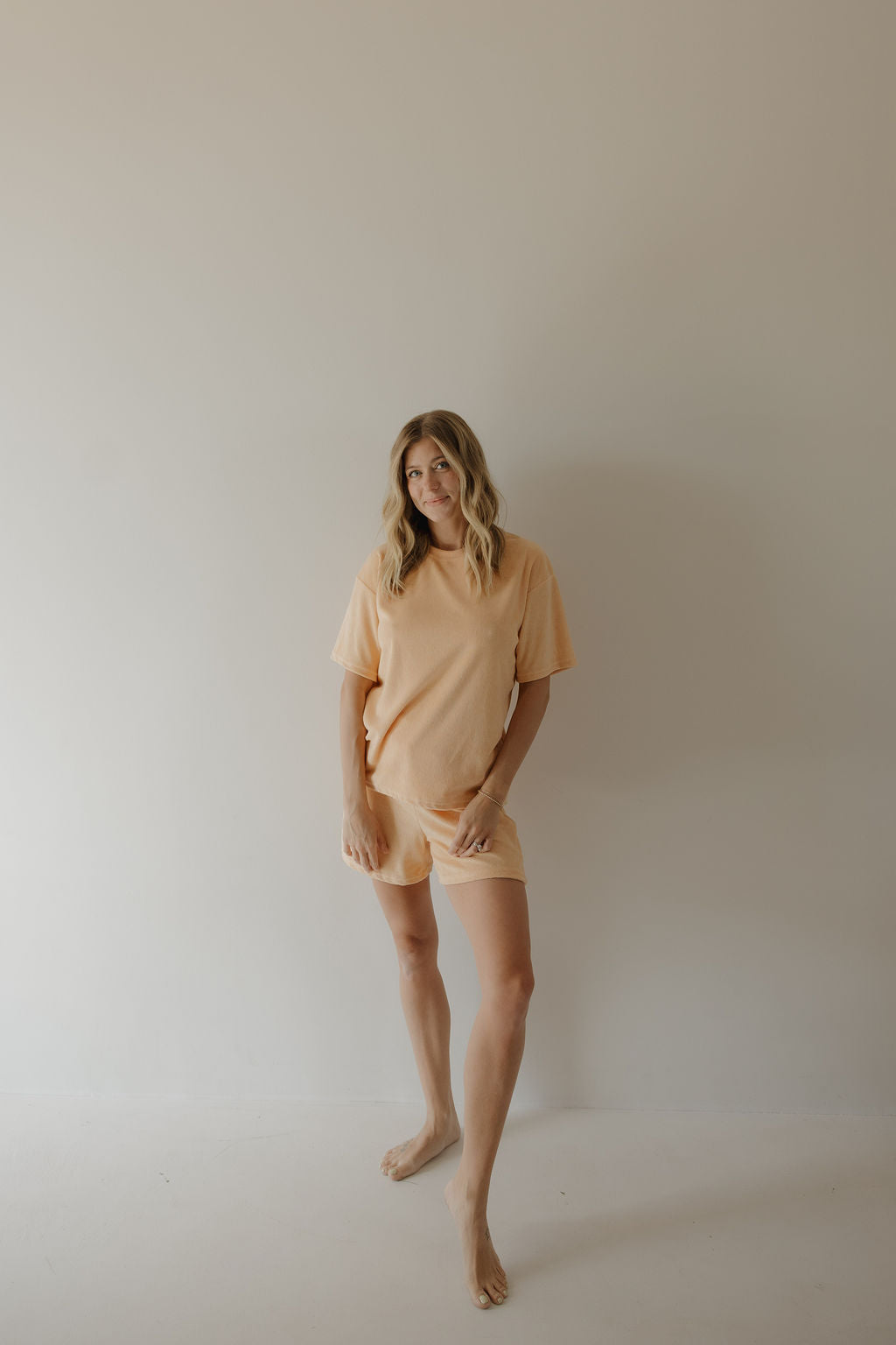 A woman with wavy blonde hair stands barefoot against a plain light-colored wall, wearing the Women's Terry Short Set in Sherbert by forever french baby. She gazes to the side with a relaxed posture, showcasing the effortless style of this set.