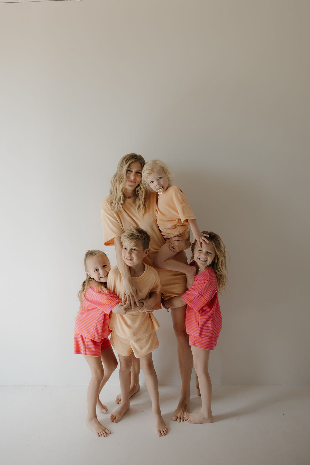 A woman stands barefoot with three smiling kids, capturing sweet family moments. The boy wears the forever french baby Child Terry Short Set in Strawberry, while two girls wear pink outfits and a baby is dressed in adorable rompers.