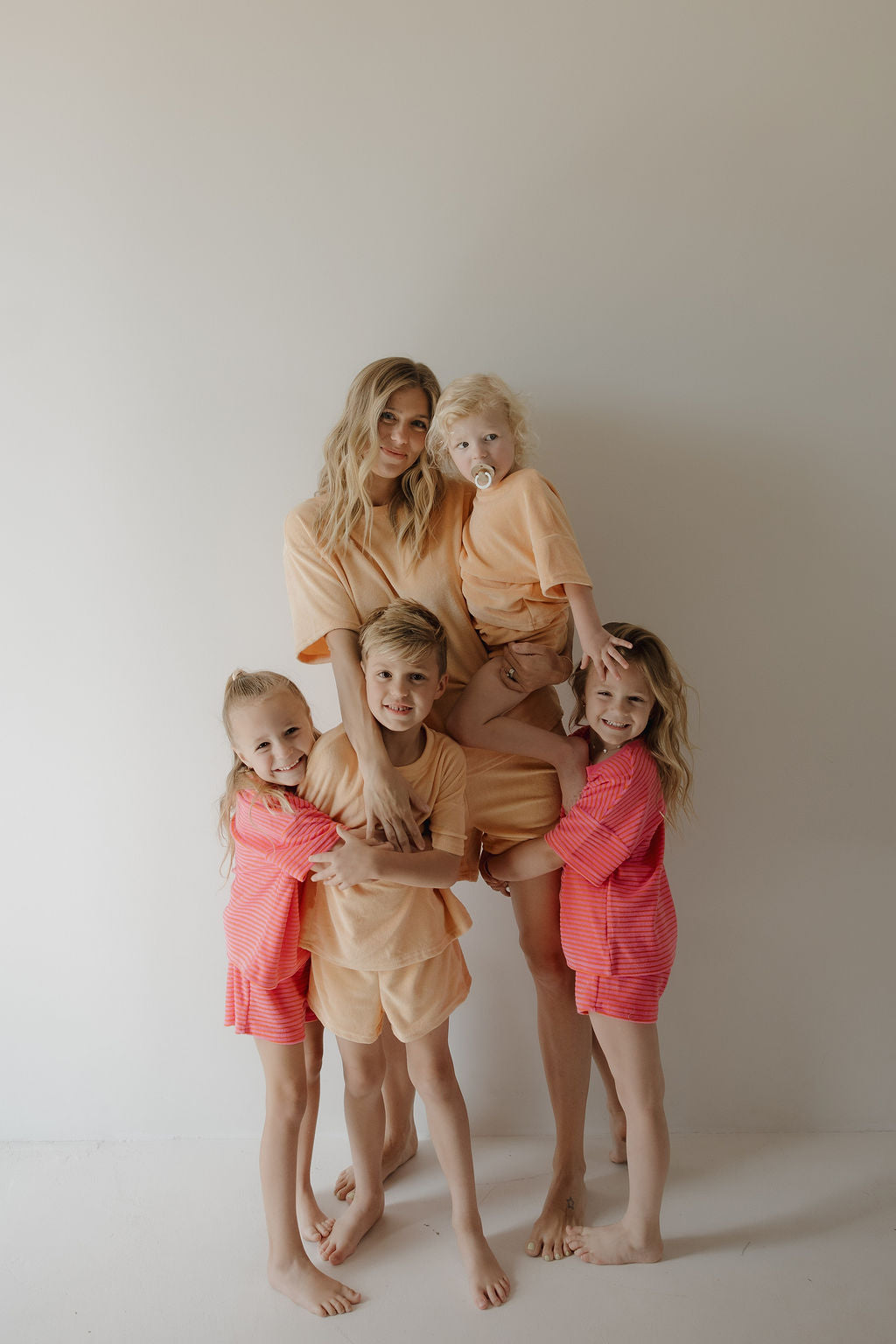 A woman in peach loungewear poses barefoot with four children, all wearing matching forever french baby Child Terry Short Set in Sherbert. They radiate warmth and joy, capturing sweet family moments against a white background.