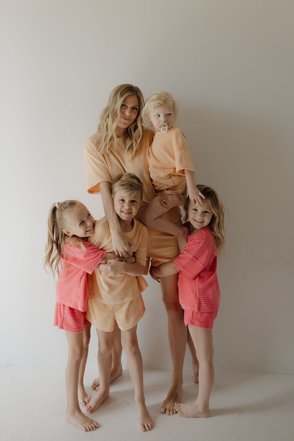 A woman stands barefoot against a plain wall in peach loungewear from forever french baby, joined by four children wearing the Child Terry Short Set | Sherbert. They smile warmly and hug, sharing affectionate family moments.