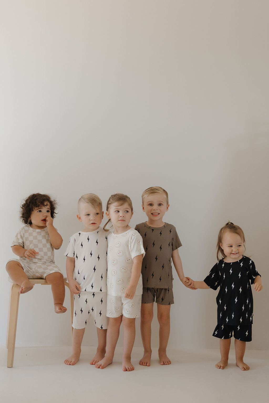 Five young children model forever french baby's Bamboo Short Set in Brown & Black Lightning Bolt. Four stand barefoot, holding hands, while one sits on a wooden stool, looking to the side in soft, breathable sleepwear against a plain background.