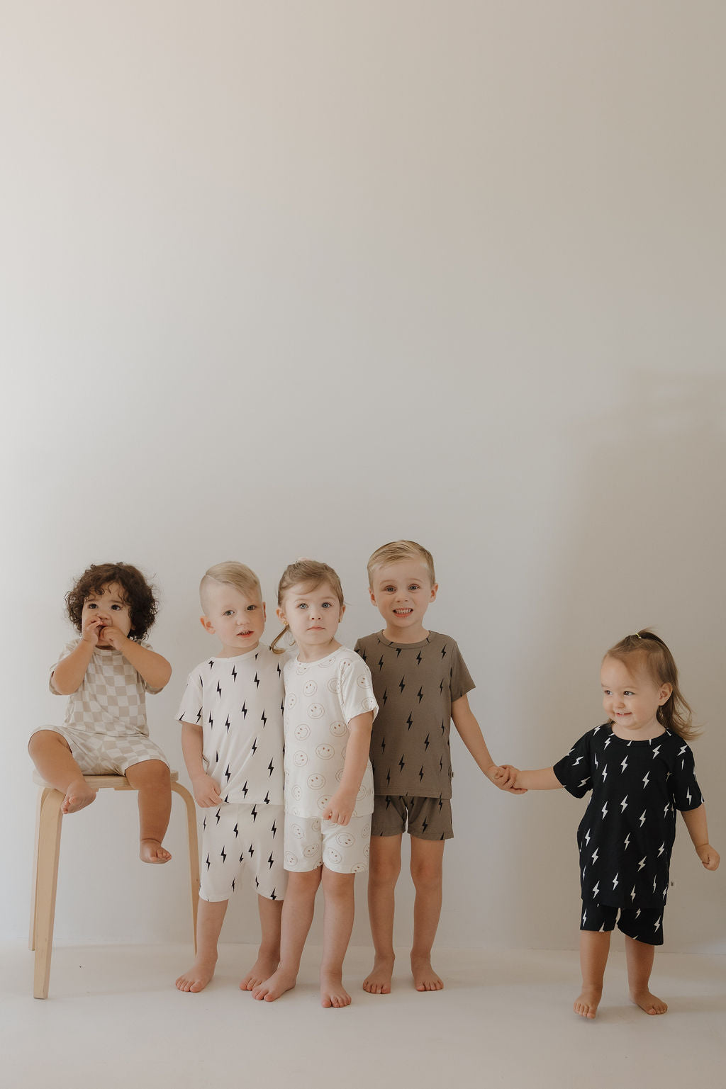 Five young children pose against a plain background. Four barefoot friends hold hands while one sits on a stool, all wearing the forever french baby Bamboo Short Set | Just Smile—matching, hypo-allergenic, breathable sleepwear in neutral patterns.