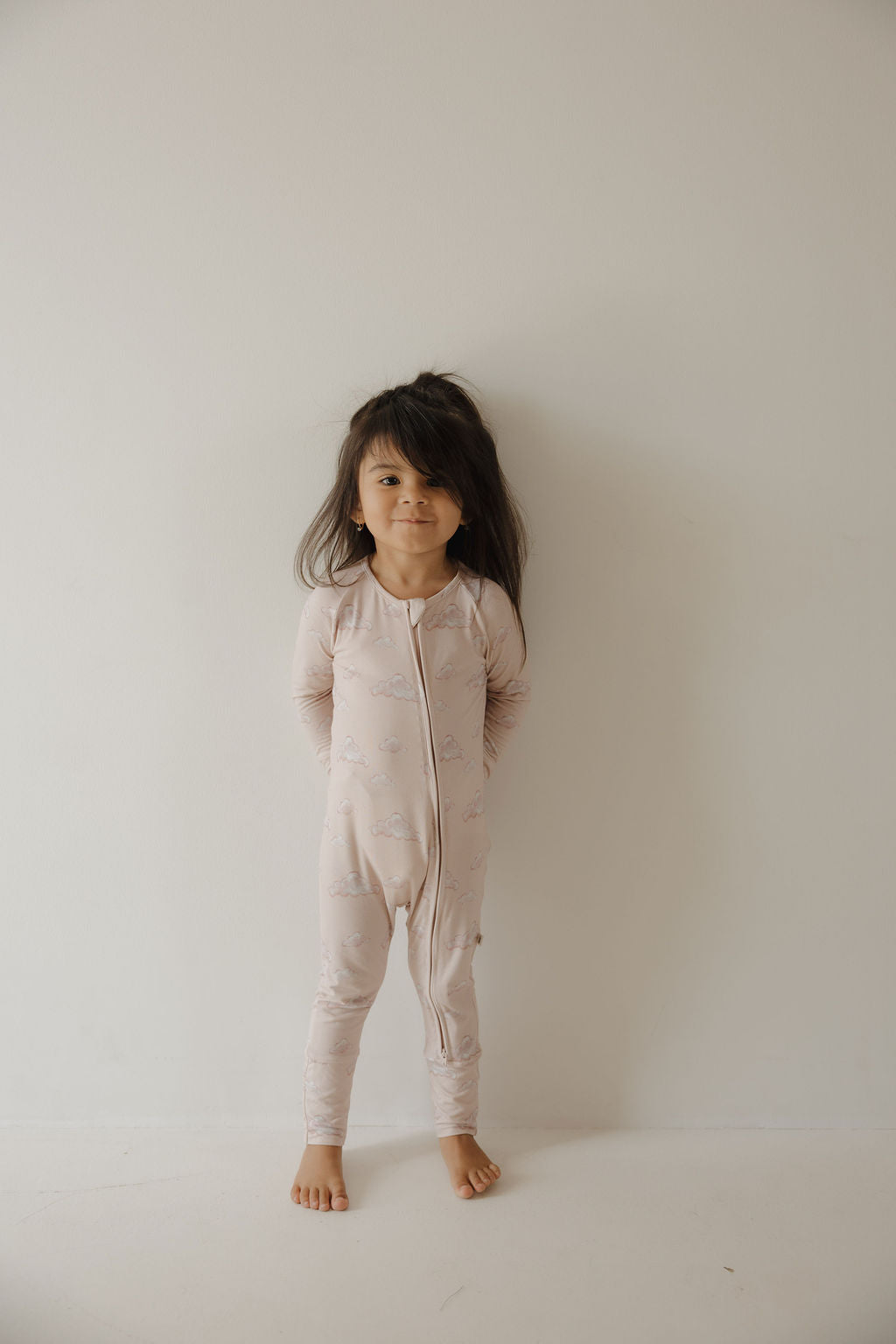 A young child with long dark hair wears Kendy 3 Bamboo Zip Pajamas in Ruby, standing barefoot against a light wall, looking at the camera with a slight smile and arms behind their back.