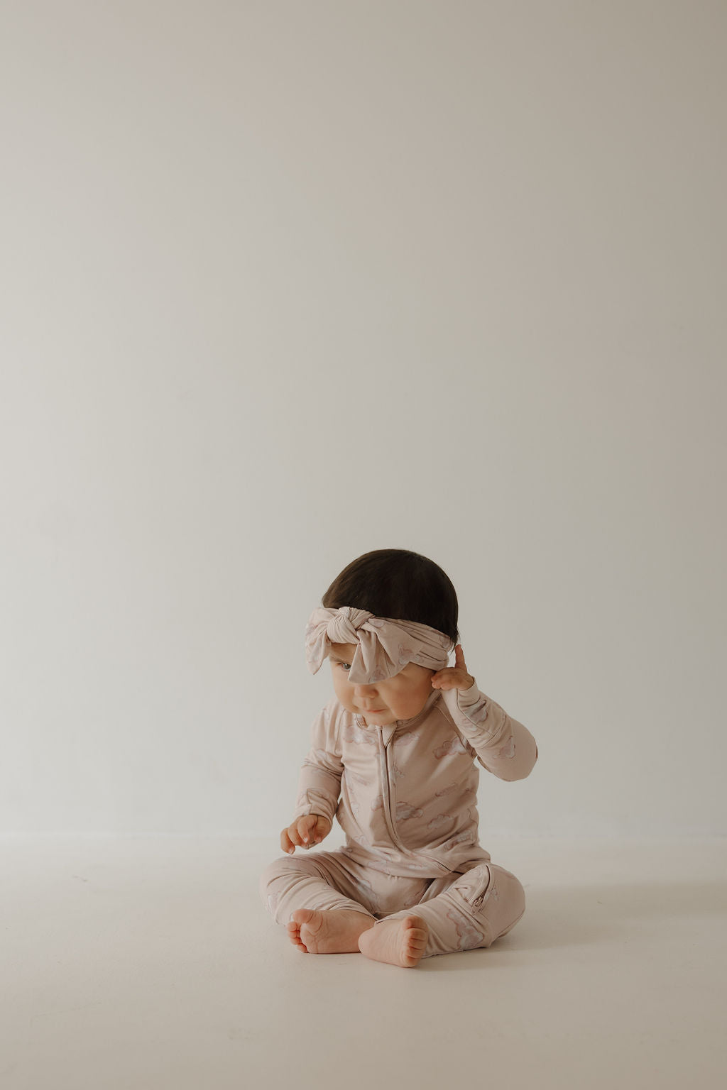 A baby in Kendy 3 Bamboo Zip Pajamas | Ruby and a matching beige headband sits barefoot on a light floor against a cream background, looking down while adjusting the headband.
