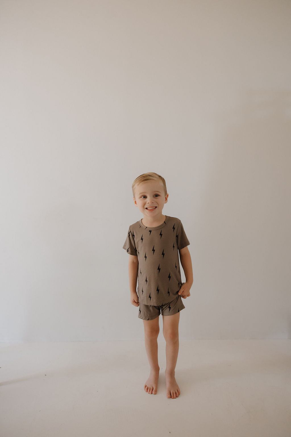 A young boy stands barefoot, smiling against a white wall, wearing the forever french baby Bamboo Short Set in Brown & Black Lightning Bolt—a hypo-allergenic matching t-shirt and shorts.