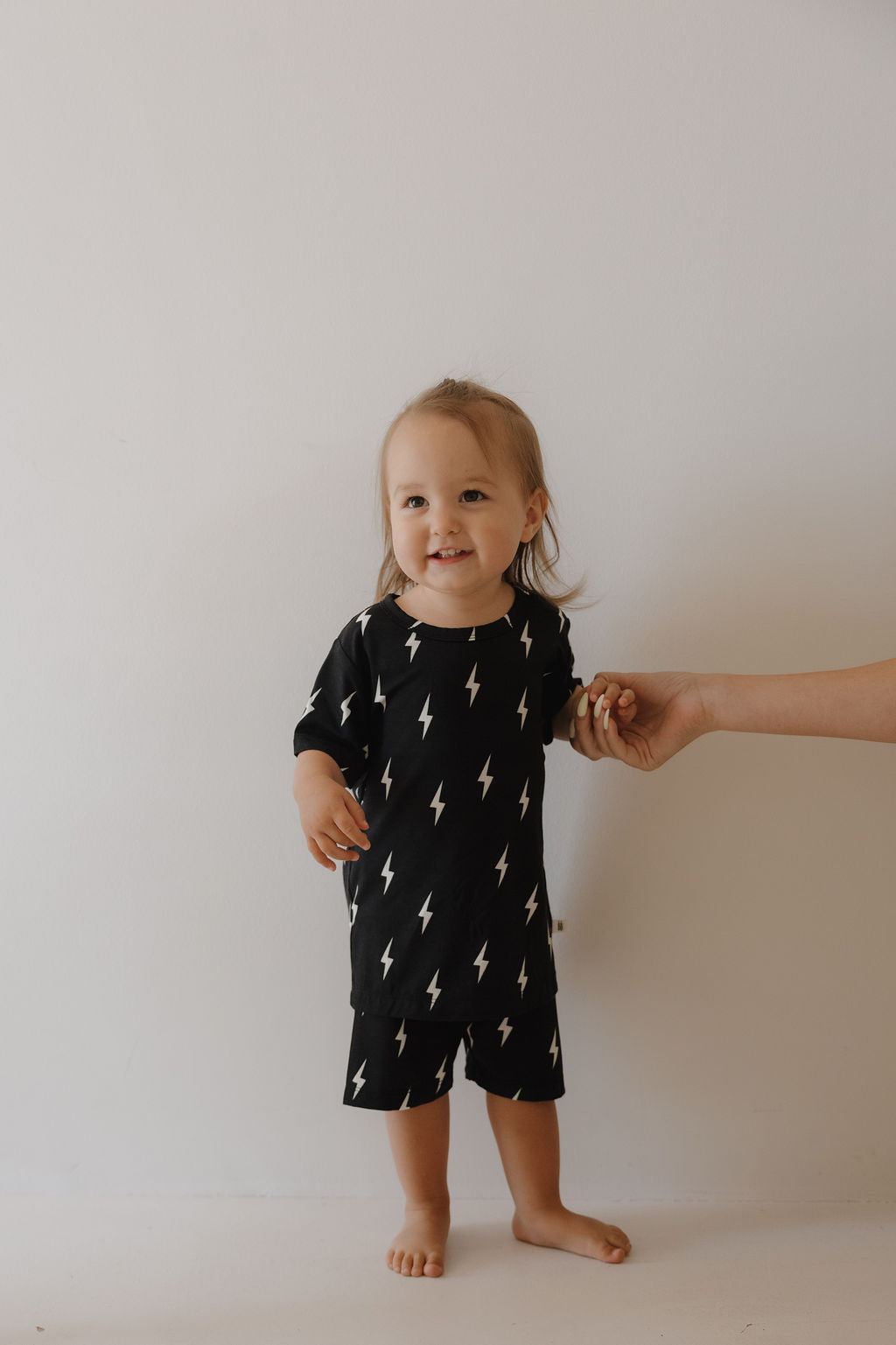 A toddler stands barefoot against a white wall, wearing forever french baby's Bamboo Short Set in Midnight Lightning Bolt, featuring a breathable design and white lightning bolt pattern, while an adult gently holds their left hand for support.