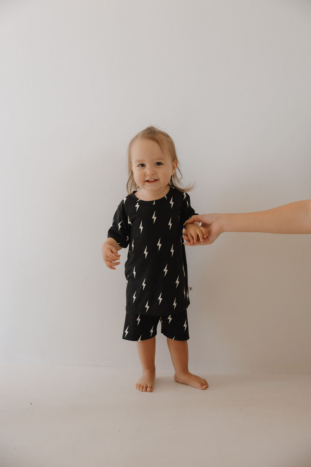 A smiling toddler wears the forever french baby Bamboo Short Set in Midnight Lightning Bolt—breathable sleepwear featuring a black outfit with white lightning bolts—standing barefoot against a plain white wall, holding an adult’s hand.