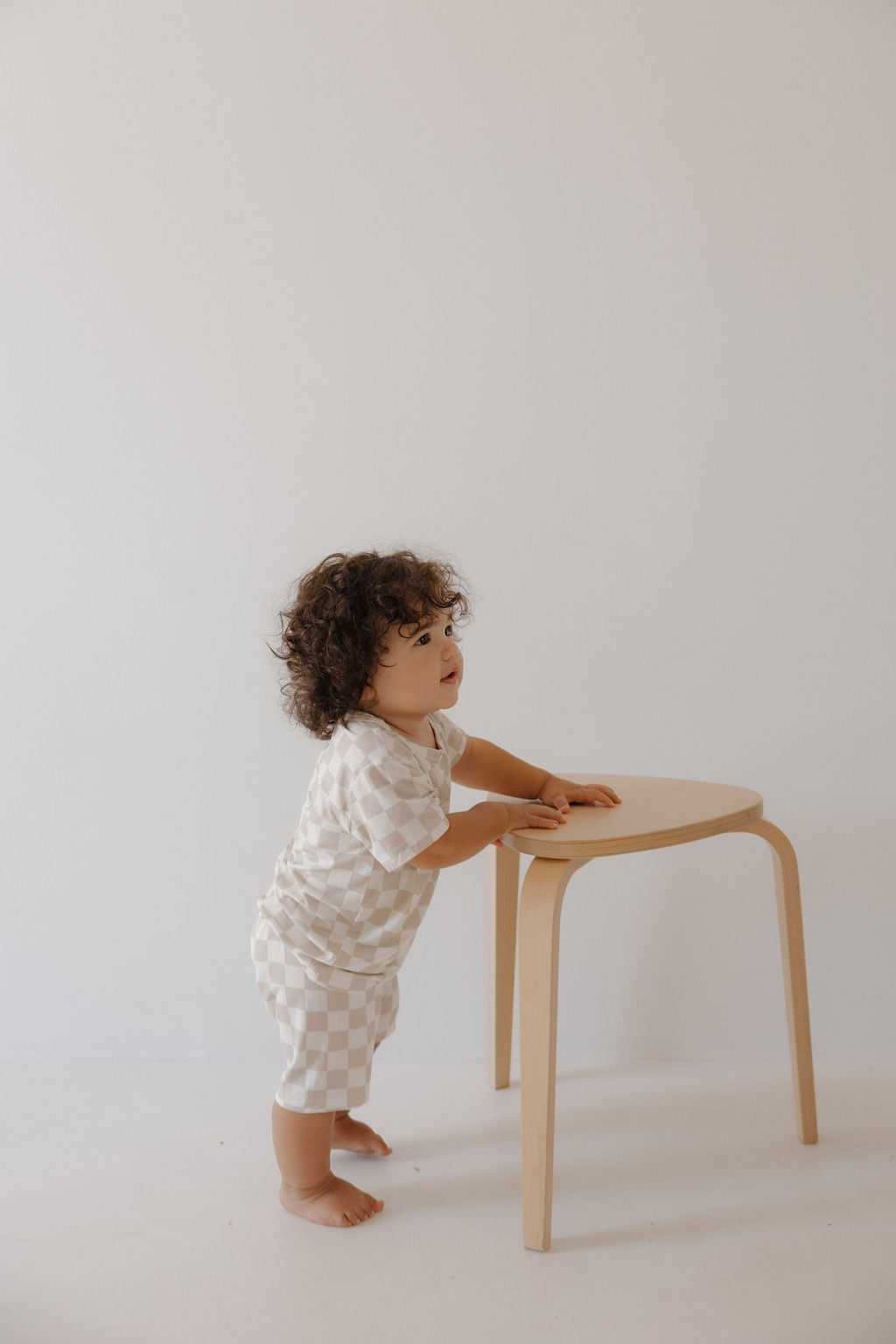 A toddler with curly hair, barefoot in the forever french baby Bamboo Short Set | Checkerboard, stands by a light wooden stool against a plain white background.