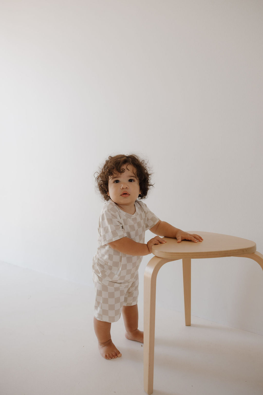 A toddler with curly hair, barefoot in a minimal white room, wears the forever french baby Bamboo Short Set | Checkerboard—breathable cream and white pajamas—standing next to a light wooden stool.
