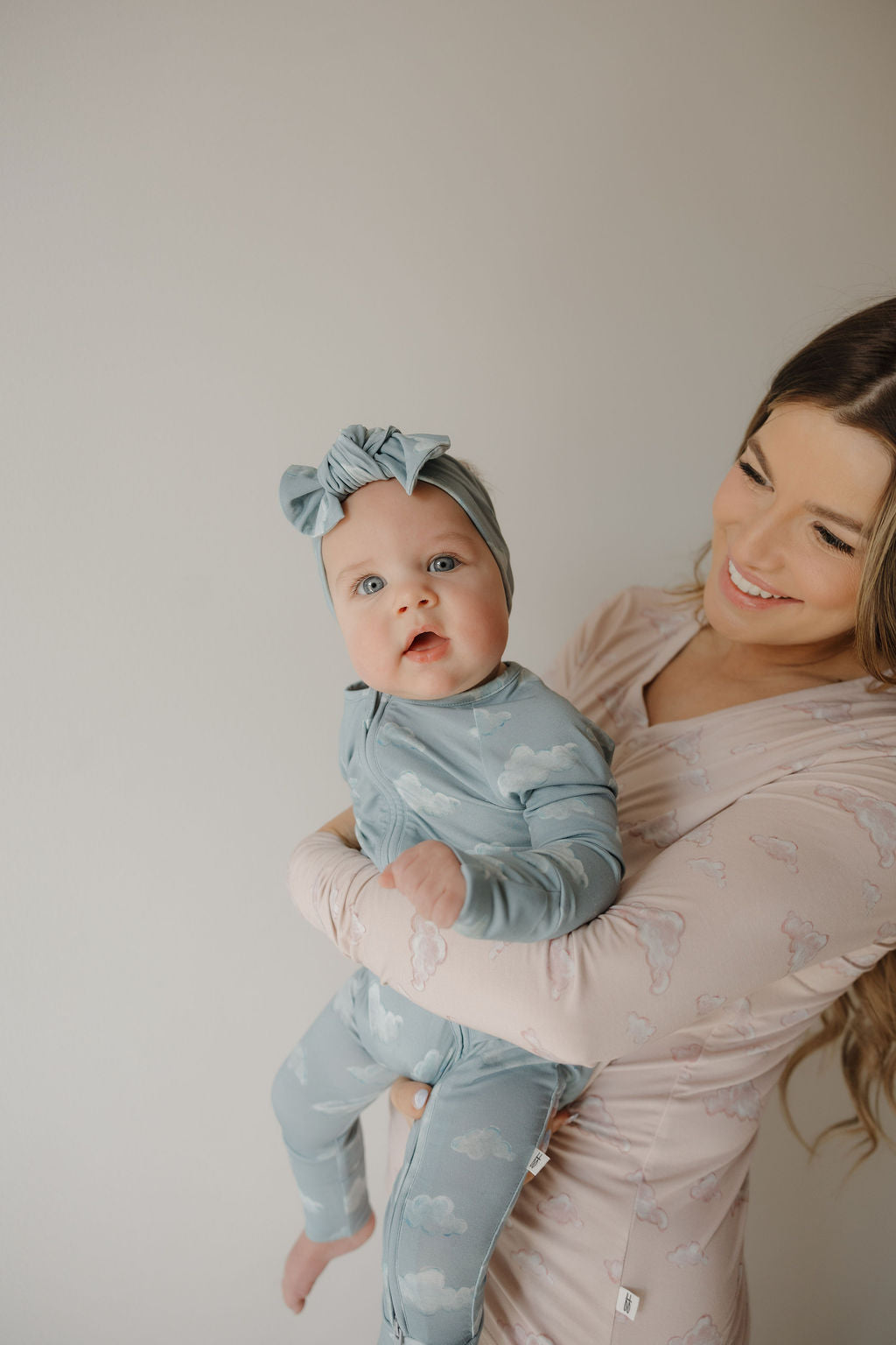 A smiling woman with long brown hair holds a baby dressed in Kendy 3’s Bamboo Zip Pajamas | Duke—a blue, cloud-print outfit with a matching headband—standing against a plain, light-colored background.