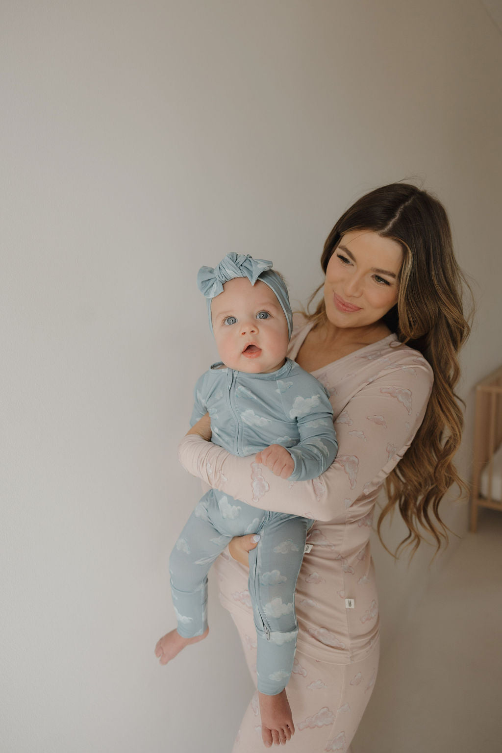 A woman with long brown hair in printed pajamas holds a baby in a blue Forever French Baby outfit and a Kendy 3 Bamboo Head Wrap from the ff x kendy collection, smiling softly indoors against a light-colored wall.