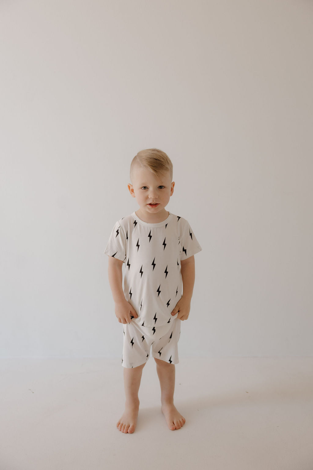 A young child stands barefoot against a plain background, wearing the forever french baby Bamboo Short Set in White & Black Lightning Bolt, with hands in the pockets.