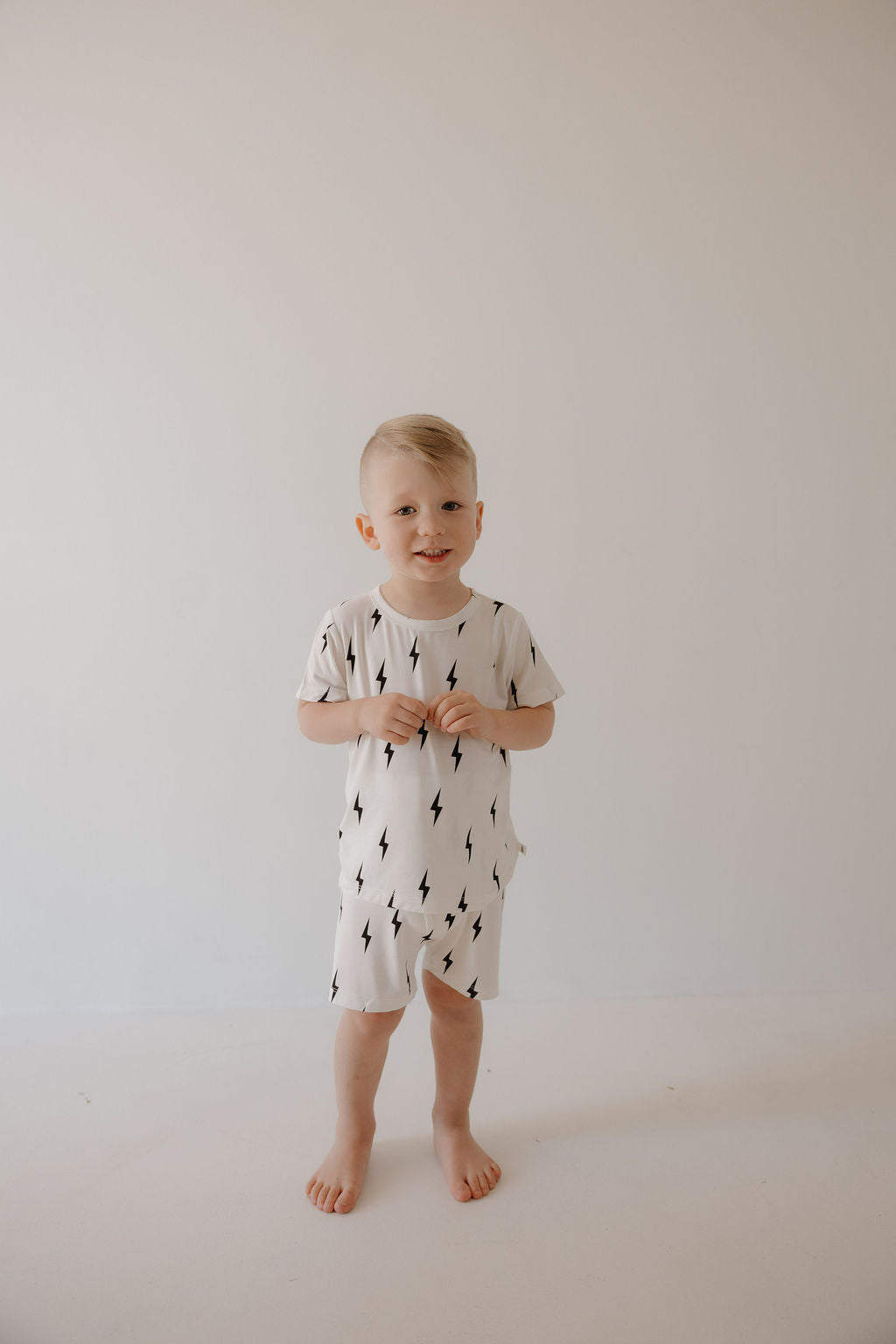 A young child with short blonde hair stands barefoot against a plain white background, wearing the forever french baby Bamboo Short Set in White & Black Lightning Bolt, hands held together at chest height.