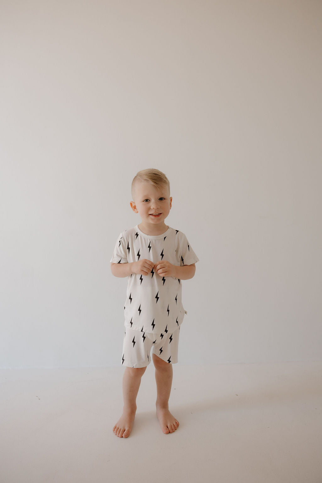A young child with short blond hair stands barefoot against a plain white background, wearing the forever french baby Bamboo Short Set in White & Black Lightning Bolt.