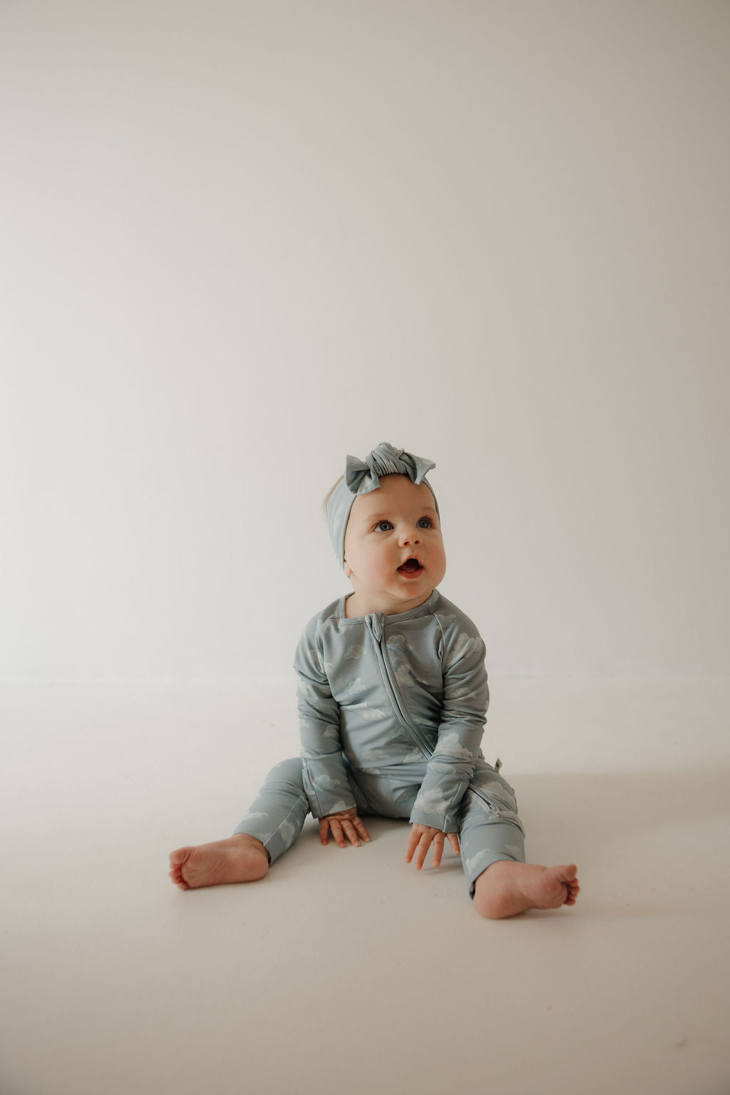 A baby in a matching light gray outfit wears the Kendy 3 Bamboo Head Wraps | ff x kendy, sitting on the floor with legs spread and a curious expression against a plain light background.