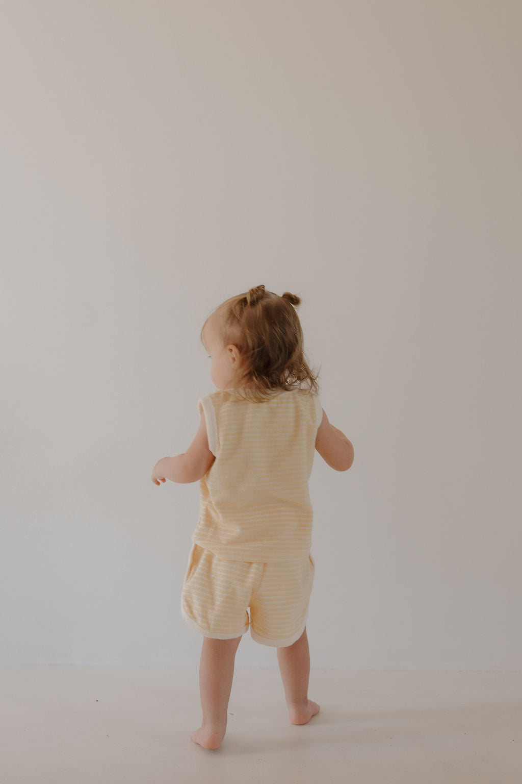 A toddler with light brown hair in a half ponytail stands barefoot, facing a plain wall, wearing the forever french baby Terry Tank Short Set | Creamsicle—featuring a sleeveless striped top and matching shorts, perfect for everyday play.