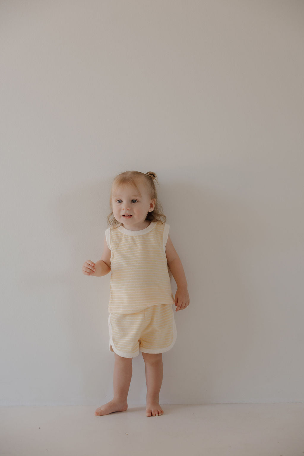 A light-haired toddler stands barefoot by a plain wall, wearing the forever french baby Terry Tank Short Set | Creamsicle—an easy, comfy choice in cream and yellow stripes for everyday fun.