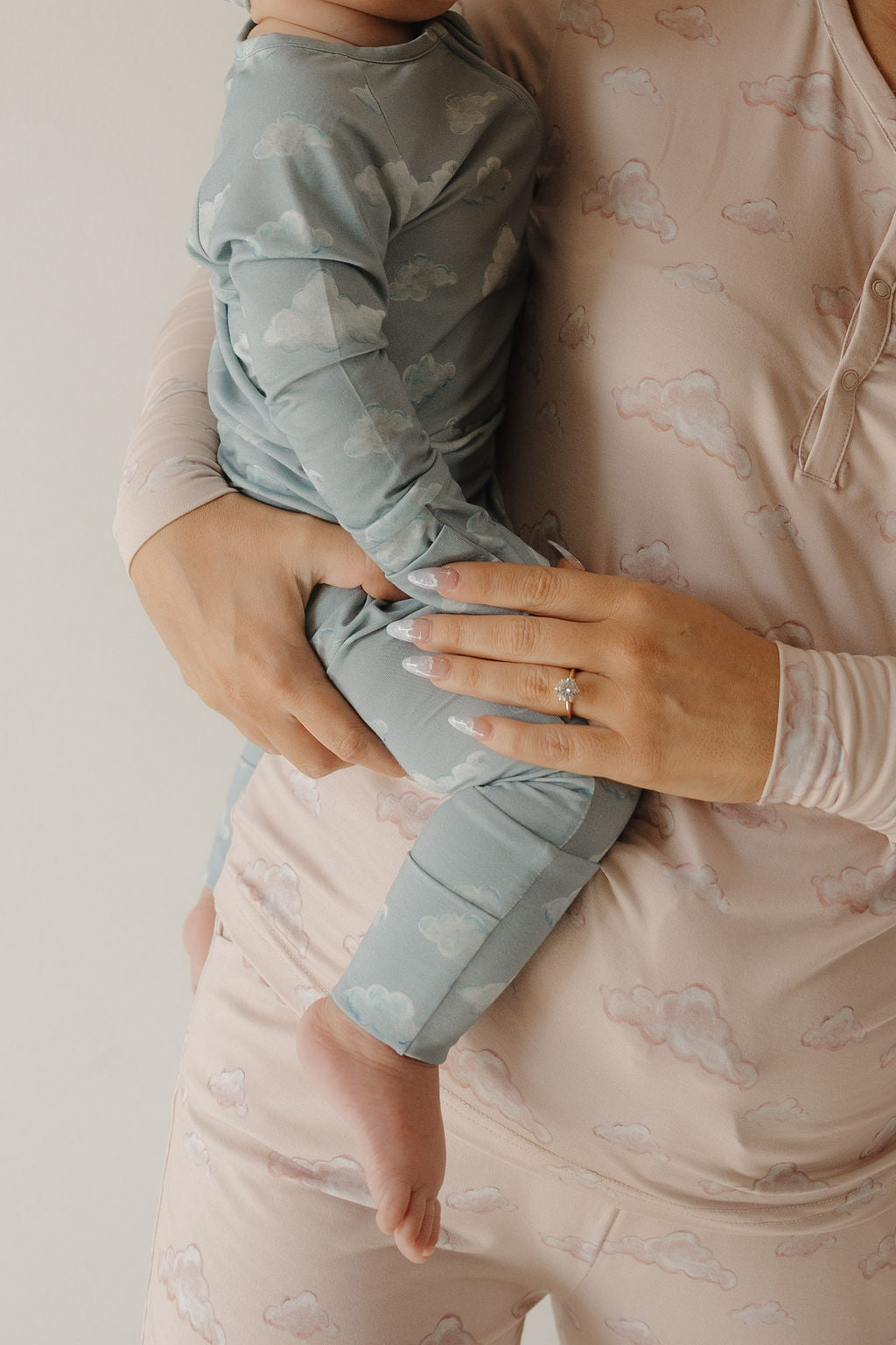 A person in pale pink cloud-print pajamas holds a baby in Kendy 3 Bamboo Zip Pajamas | Duke, featuring a blue cloud print. The person's ringed hand gently supports the baby as both wear breathable, soft bamboo fabric outfits.