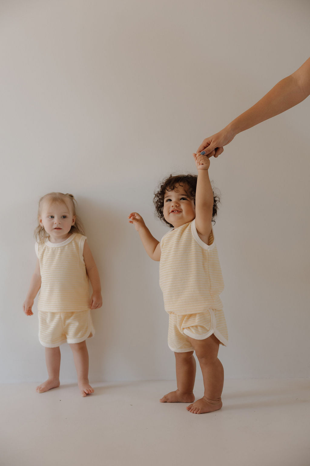 Two young children in matching forever french baby Terry Tank Short Set | Creamsicle outfits stand against a light background. One smiles, holding an adult’s hand, while the other stands nearby, ready for everyday adventures.