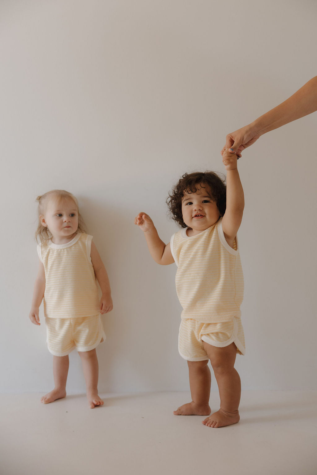Two young children in matching Forever French Baby Terry Tank Short Set in Creamsicle stand against a plain background. One holds an adult’s hand, while the other stands nearby—ready for everyday adventures.