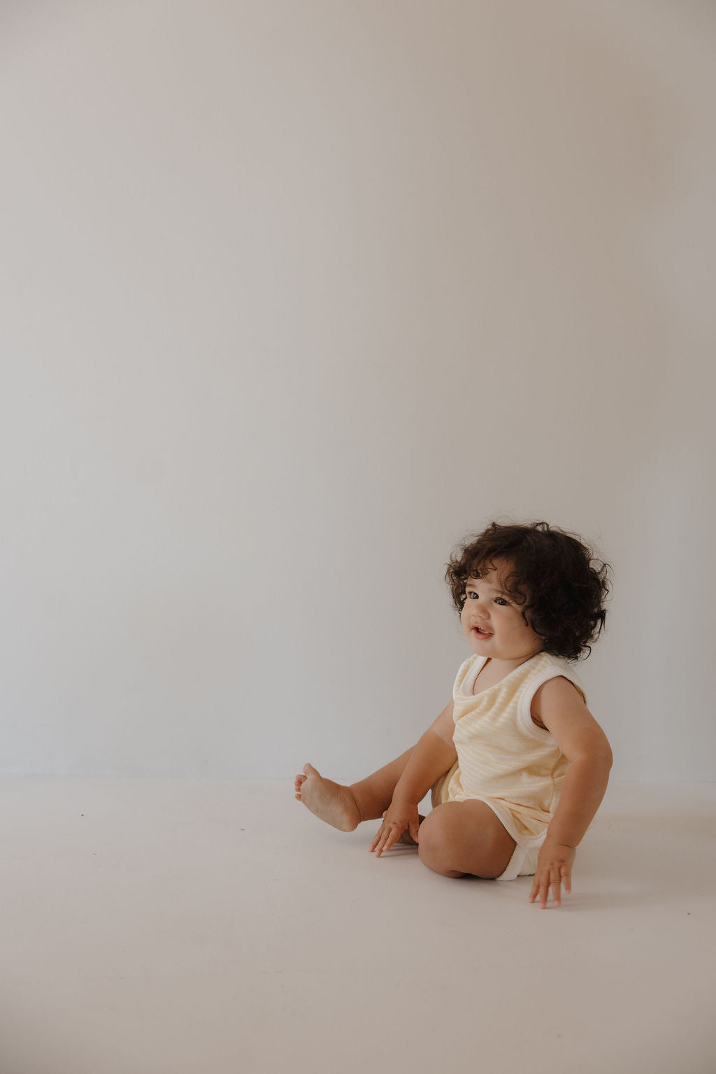 A young child with curly dark hair sits against a light wall, wearing the forever french baby Terry Tank Short Set in Creamsicle and gazing to the side—perfectly dressed for everyday adventures.