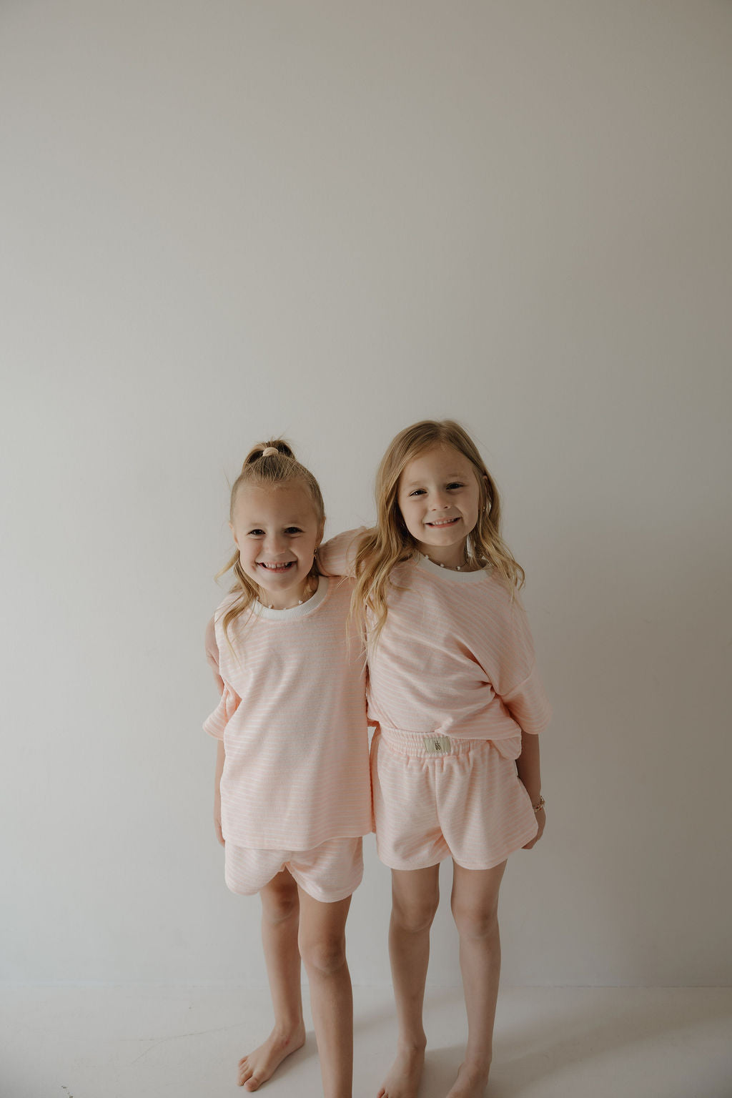 Two young girls with blonde hair stand barefoot against a plain light background, smiling and wearing matching forever french baby Child Terry Short Set in Cotton Candy, capturing the joy of family moments.