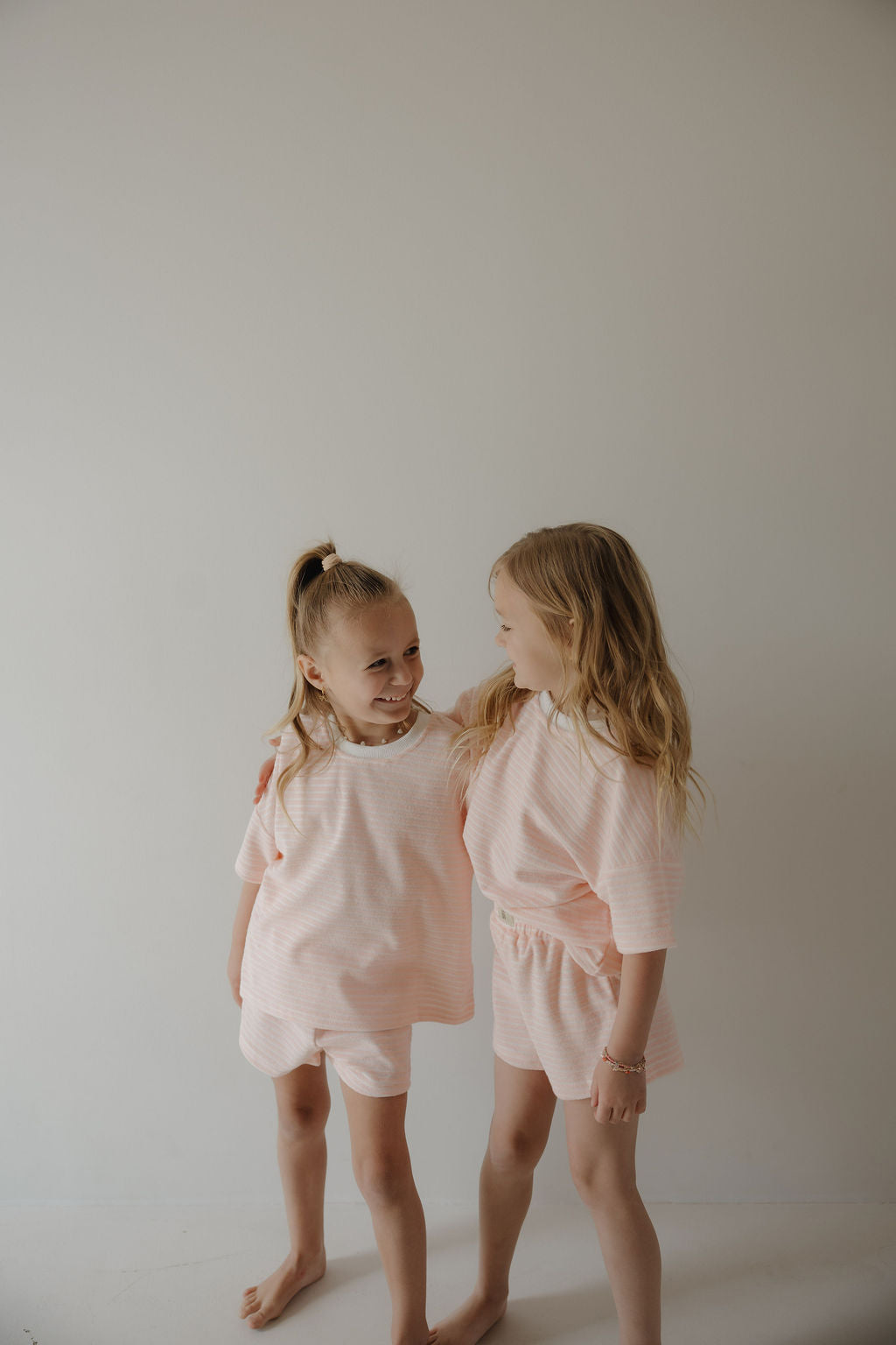Two young girls smile warmly, standing close in matching Forever French Baby Child Terry Short Sets in Cotton Candy, one with her arm around the other, capturing sweet family moments against a simple light background.