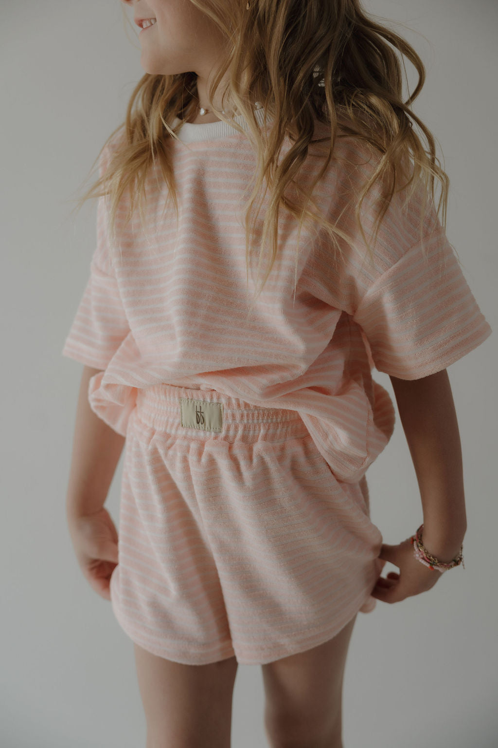 A young child with wavy light brown hair smiles while wearing the forever french baby Child Terry Short Set in Cotton Candy, featuring light pink and white stripes—perfect for capturing sweet family moments against a neutral background.