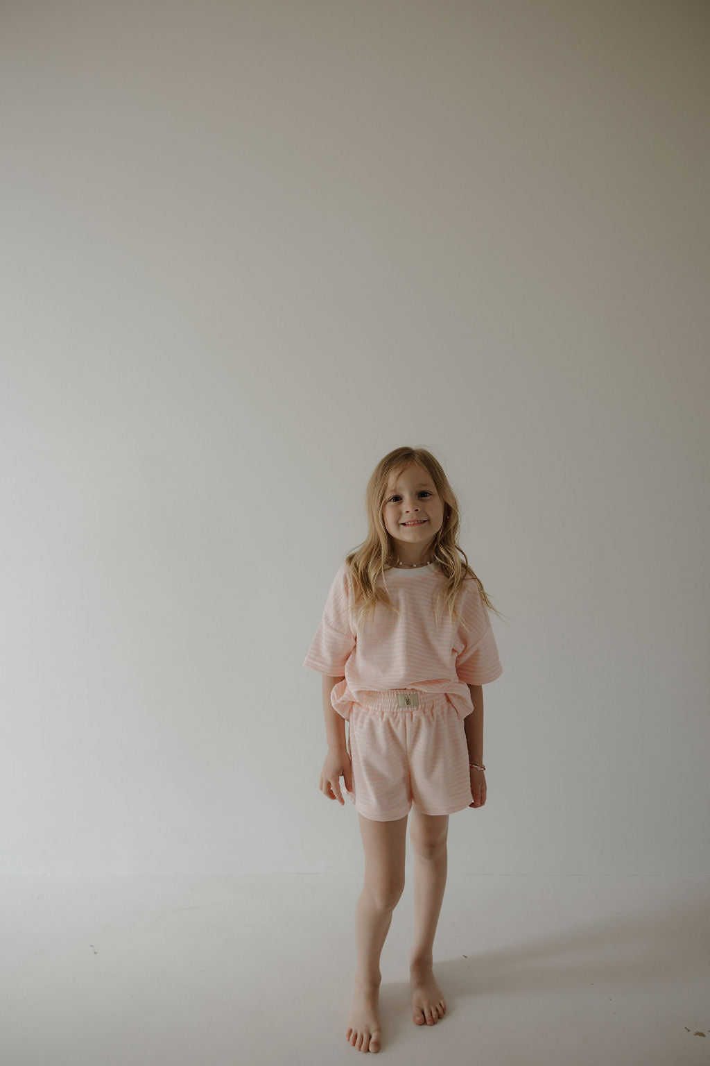 A young girl with long blonde hair stands barefoot against a light wall, smiling softly in a forever french baby Child Terry Short Set | Cotton Candy, capturing sweet family moments.