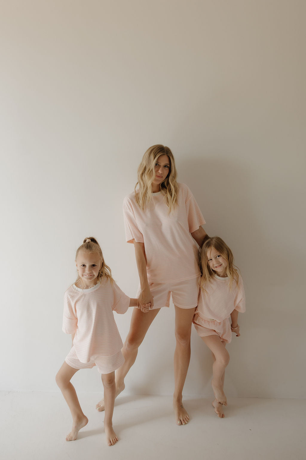 A woman and two young girls stand barefoot by a light wall, all in matching forever french baby Child Terry Short Set in Cotton Candy, sharing relaxed family moments as the woman holds each girl’s hand.