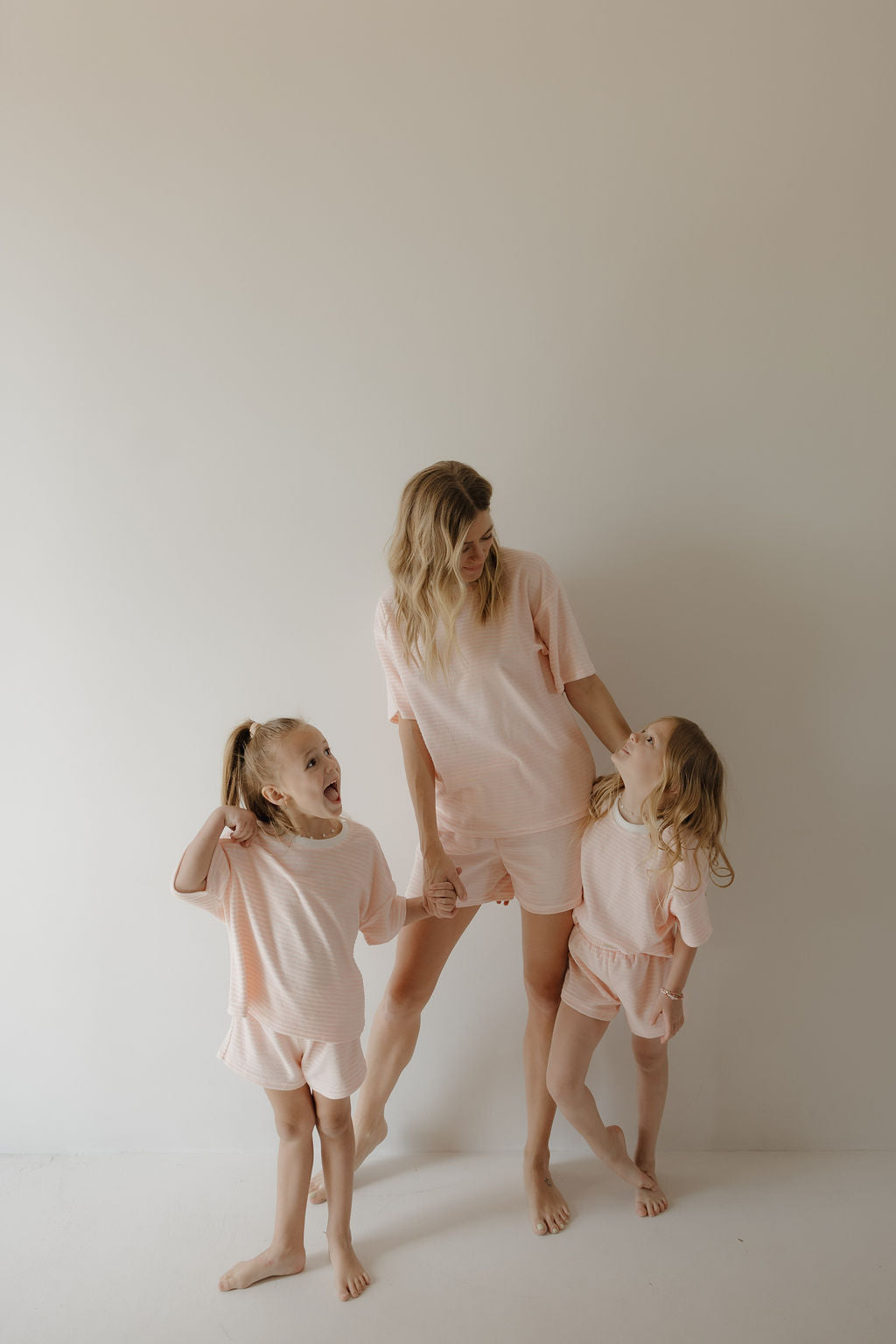 A woman and two girls, barefoot in matching forever french baby Child Terry Short Set | Cotton Candy, stand smiling and holding hands against a plain wall—capturing sweet family moments in our cozy loungewear.