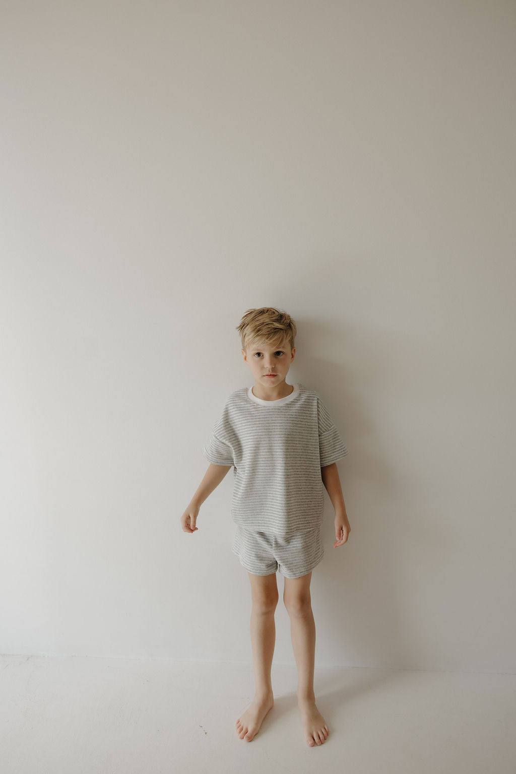 A young boy with blonde hair stands barefoot against a white wall, wearing the forever french baby Child Terry Short Set | Grey Stripe, looking at the camera with a neutral expression—ideal for family photos.