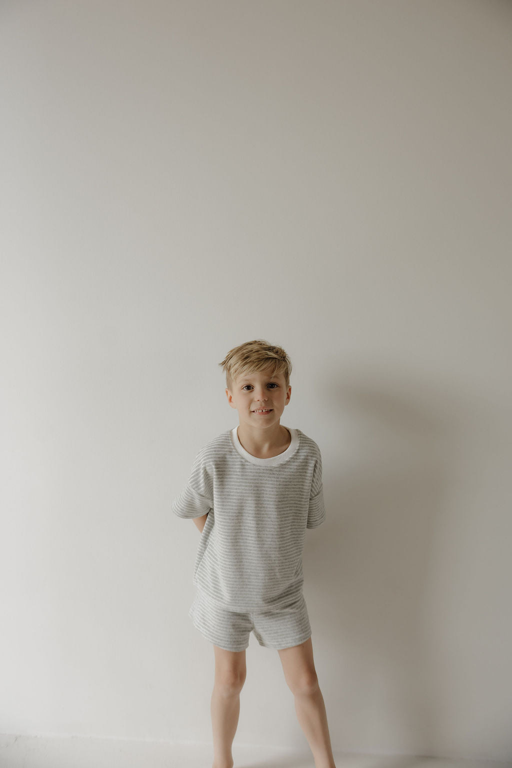 A young boy with blonde hair stands barefoot against a white wall, wearing the forever french baby Child Terry Short Set | Grey Stripe, hands behind his back as he looks at the camera, capturing a simple family moment.