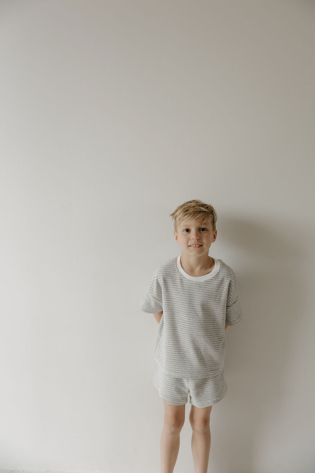 A young boy with blonde hair stands before a plain white wall, wearing the forever french baby Child Terry Short Set in Grey Stripe—hands behind his back and a slight smile, capturing simple family moments.