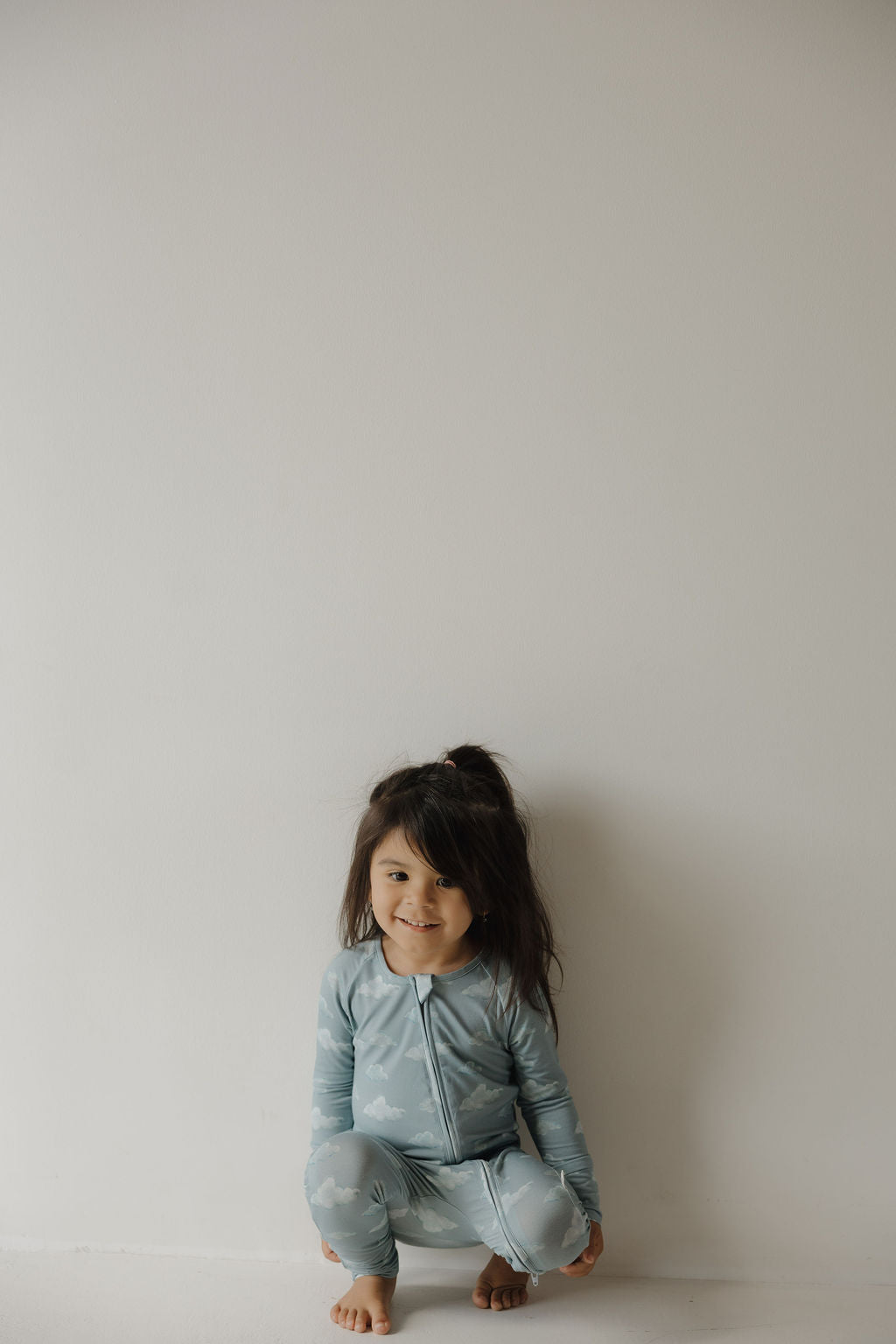 A young child with long dark hair squats barefoot by a white wall, smiling softly in Kendy 3's Bamboo Zip Pajamas | Duke—light blue pajamas with a cloud pattern and breathable fabric for all-day comfort.