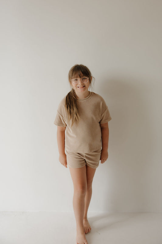 A young girl with long hair and glasses stands barefoot against a white wall, wearing the forever french baby Child Terry Short Set in Tan, smiling softly and capturing simple family moments.