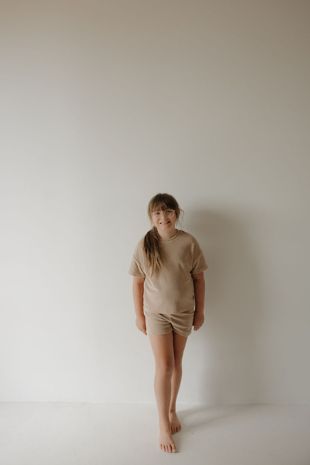 A young girl with long hair stands barefoot against a plain white wall, wearing the forever french baby Child Terry Short Set in Tan. She looks relaxed with her arms by her sides, reflecting the simplicity of family moments.