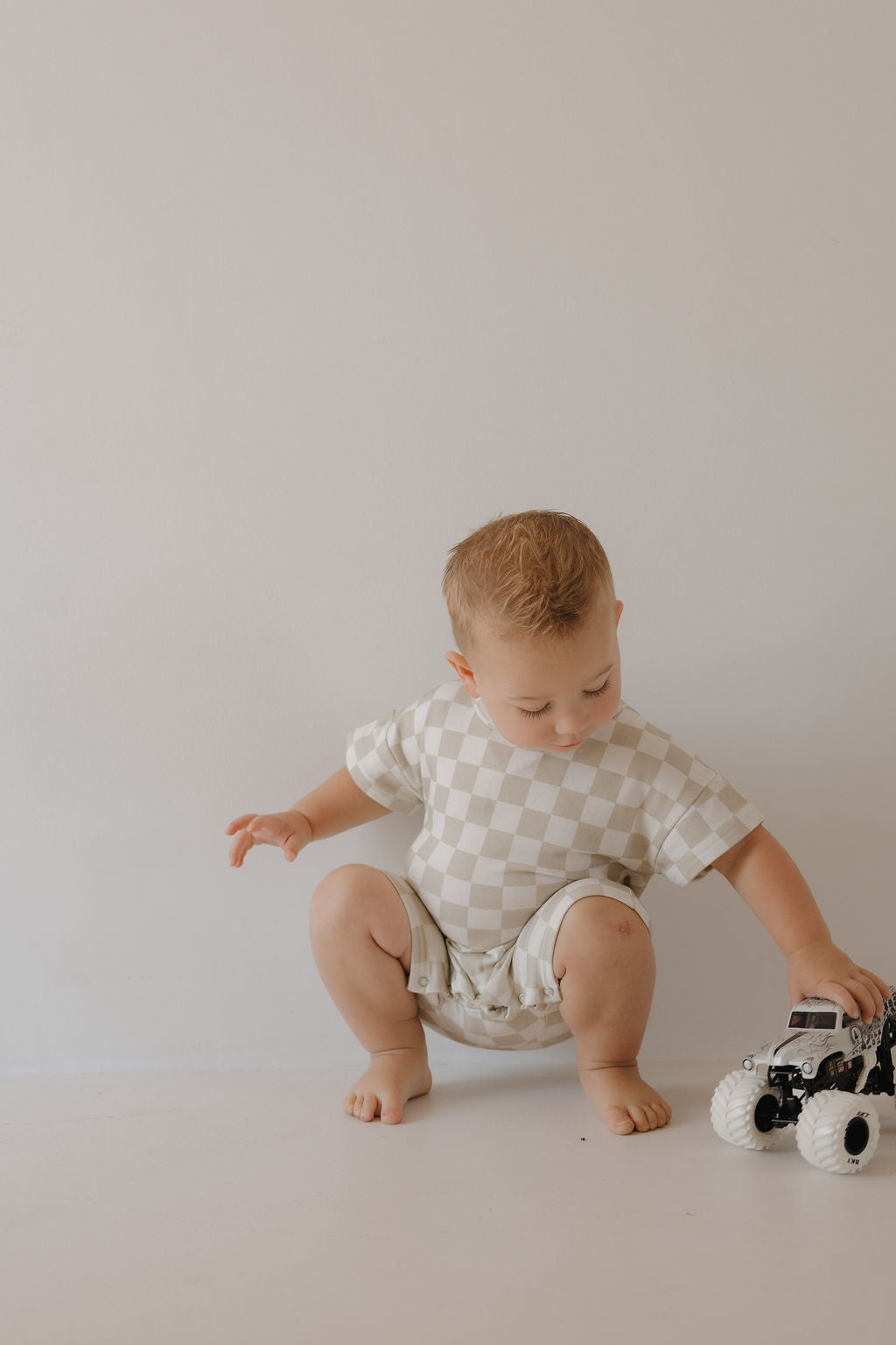 A toddler with short blond hair wears the forever french baby French Terry Romper | Checkerboard while playing with a black and white toy truck on a plain light background. Fits newborns to 24 months.