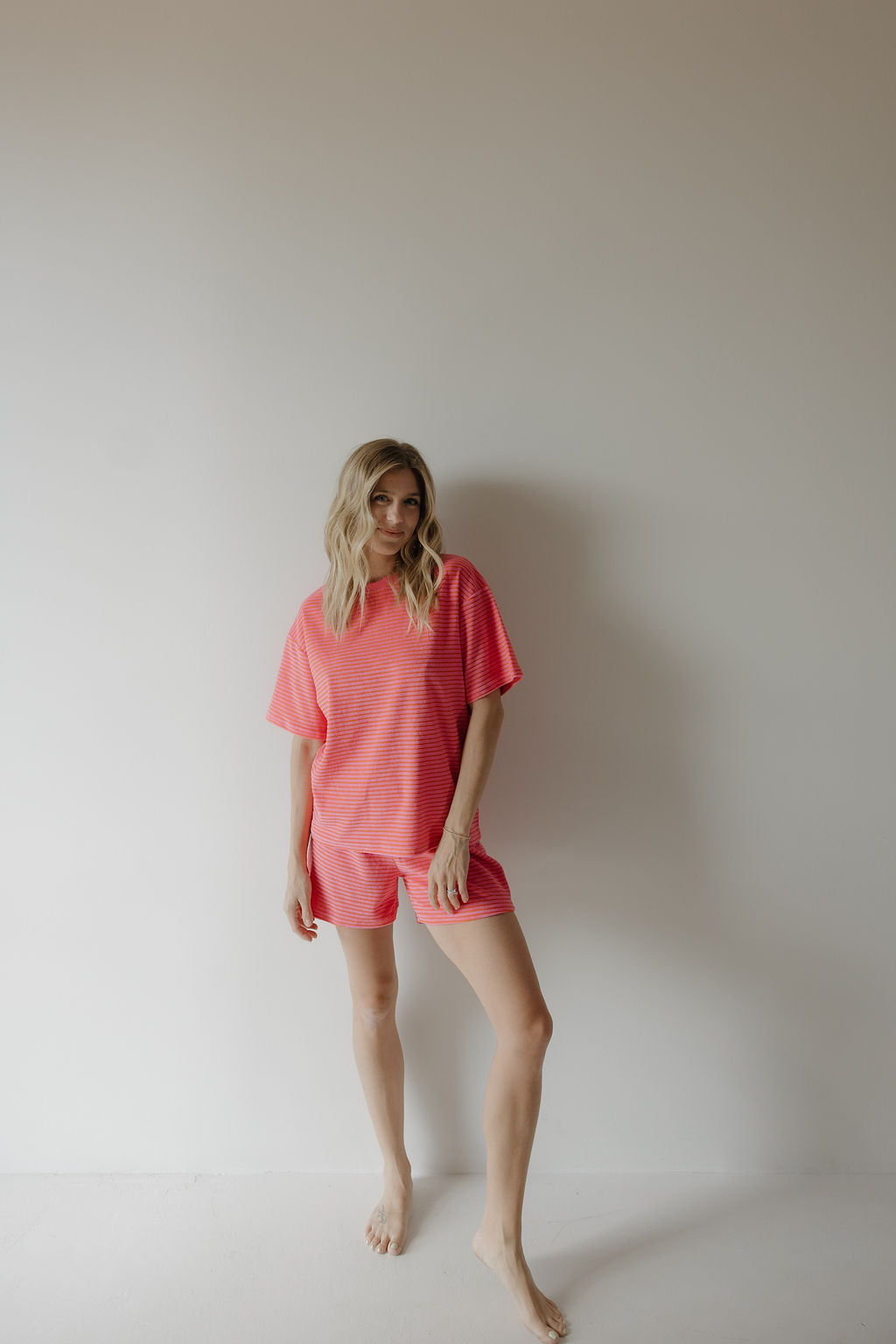 A barefoot woman with blonde hair stands against a plain white wall, wearing the forever french baby Women's Terry Short Set in Strawberry. She gazes at the camera, showcasing effortless style and comfort.