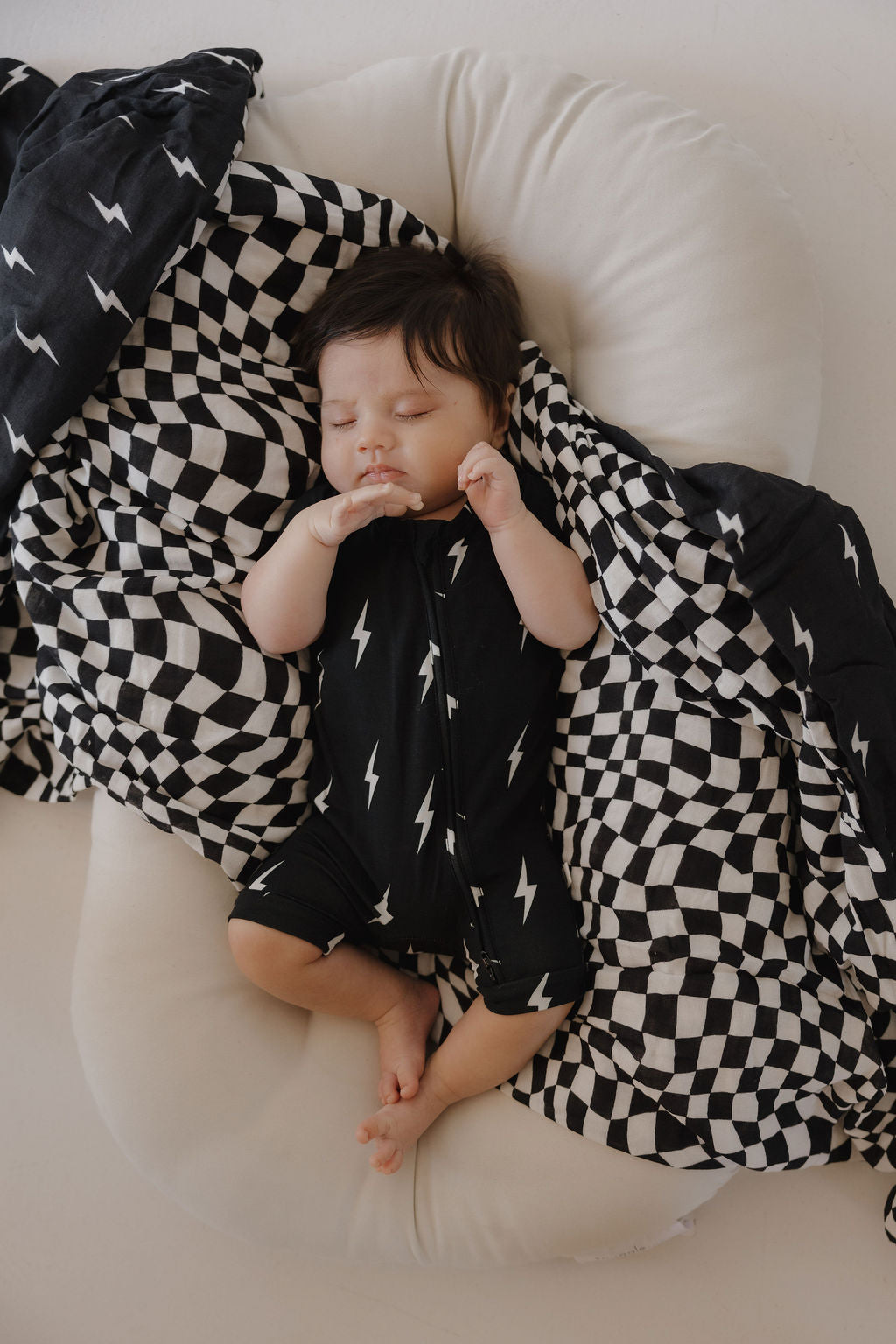 A baby sleeps peacefully on a white cushion, dressed in the forever french baby Bamboo Zip Shortie | Midnight Lightning Bolt, surrounded by black-and-white checkered and lightning-patterned blankets.