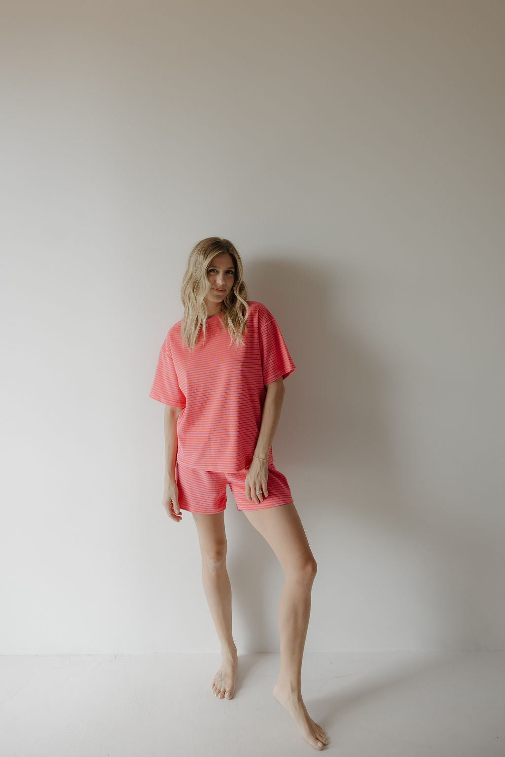 A woman with long blonde hair stands barefoot against a plain white wall, wearing the cozy Women's Terry Short Set in Strawberry from forever french baby—perfect for relaxing or enjoying special family moments.