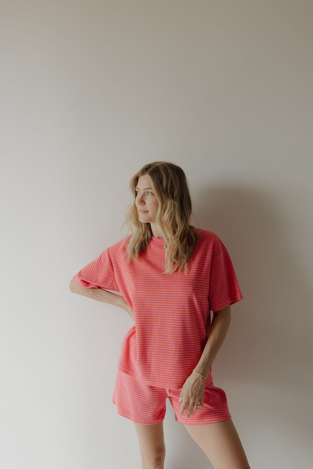 A woman with wavy blonde hair stands against a light wall, wearing the Women's Terry Short Set in Strawberry from forever french baby. She looks to the side with one hand on her hip.