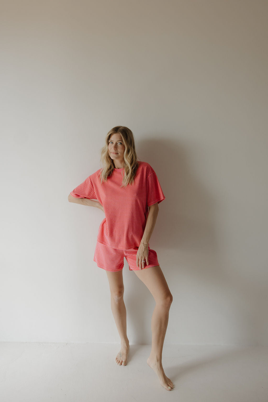 A blonde woman stands barefoot against a light wall, wearing the Women's Terry Short Set in Strawberry by forever french baby—her relaxed, pink-striped top and shorts radiate comfort as she looks sideways with one hand on her hip.