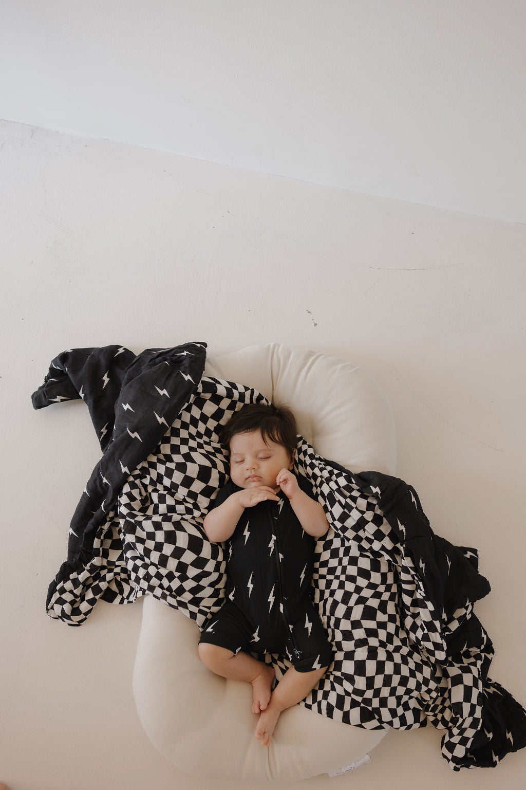 A baby sleeps on a white cushion, wrapped in a black and white checkered blanket with a matching lightning bolt design. The baby wears the forever french baby Bamboo Zip Shortie | Midnight Lightning Bolt, featuring a stylish lightning bolt print.