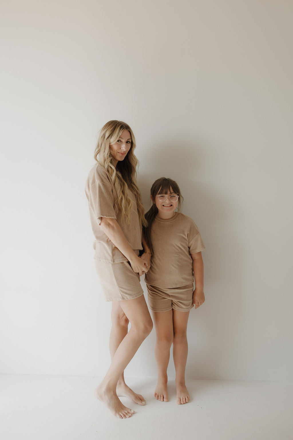 A woman and a young girl stand barefoot against a white wall, both wearing the forever french baby Child Terry Short Set in Tan. Smiling softly and holding hands, they capture the warmth of family moments.