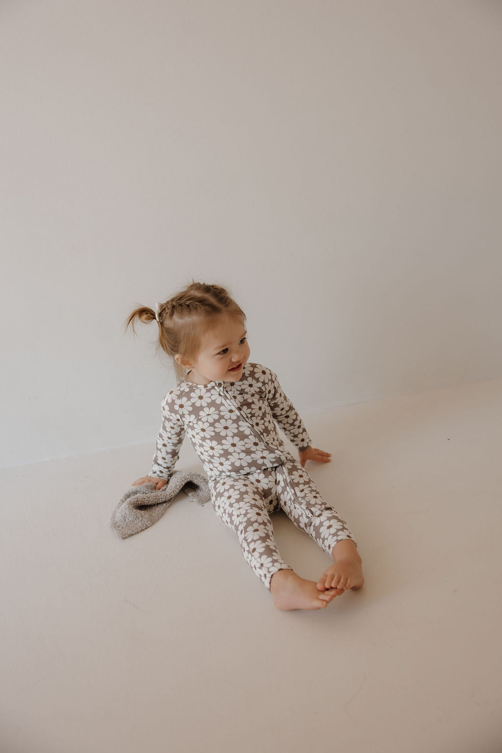 A young child with light brown hair in pigtails sits barefoot on a light floor, smiling in forever french baby's Bamboo Zip Pajamas | Darlin'. A plush toy beside her highlights the cozy, breathable comfort of these floral-patterned pajamas.